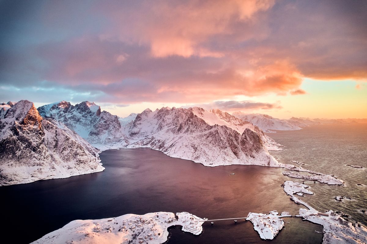 Pink sunrise sky over snowy mountains and the dark fjord around Reine, seen from a drone